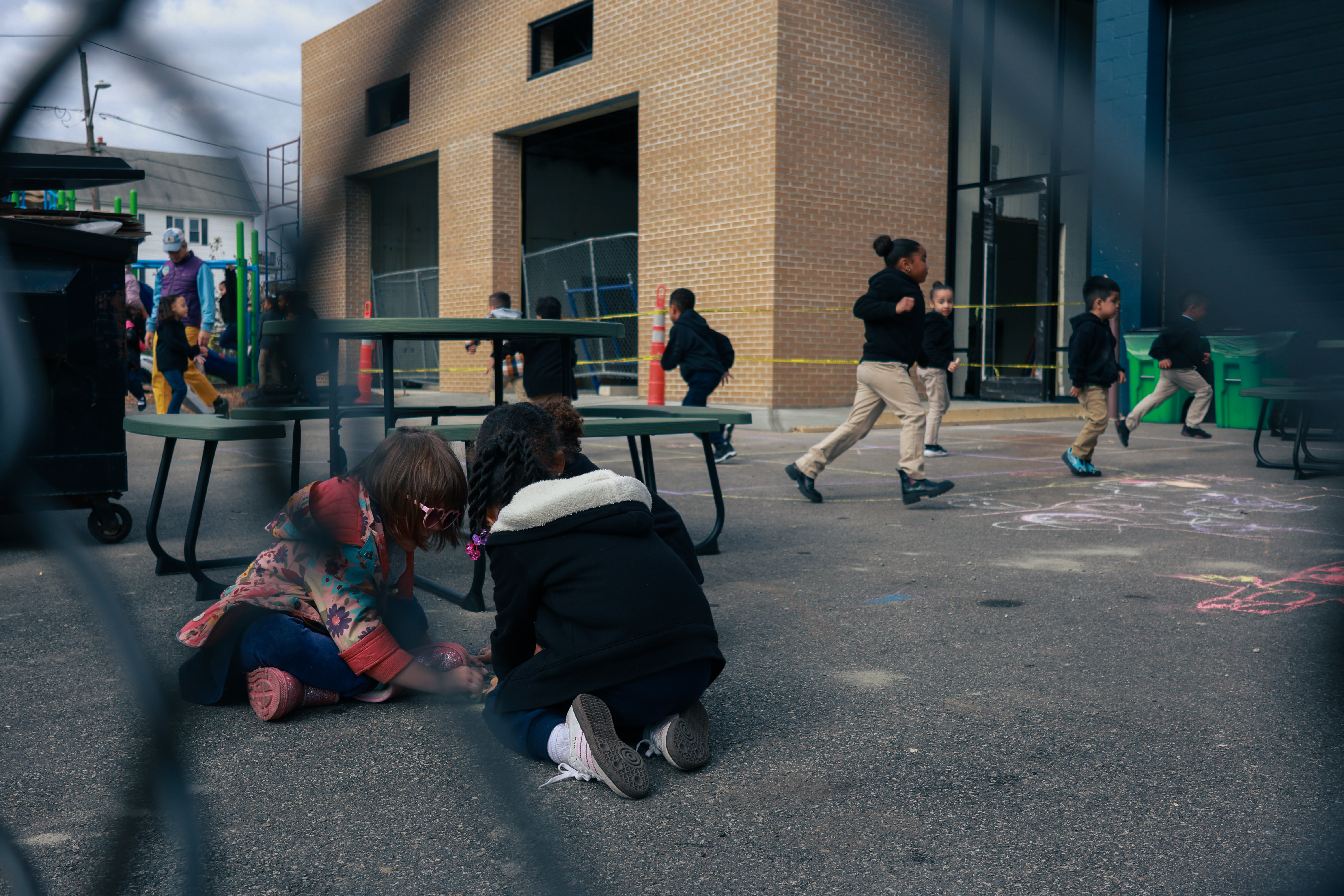 Through the links of a fence, three students can be seen huddled together drawing with sidewalk chalk. Others are running in the background.